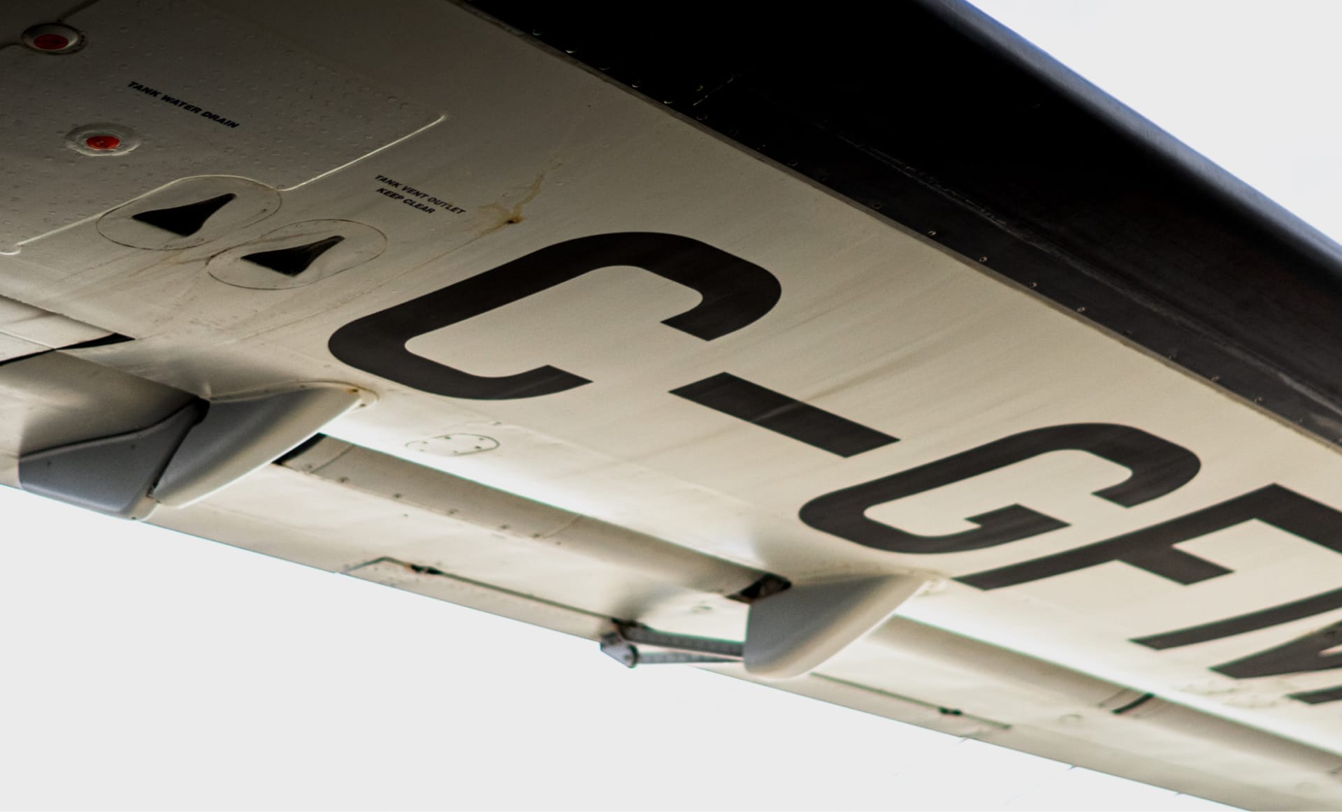 Close-up view of an aircraft wing from below, showing part of its registration number C-GF painted in black on the white surface, with visible hinges, panels, and vents.