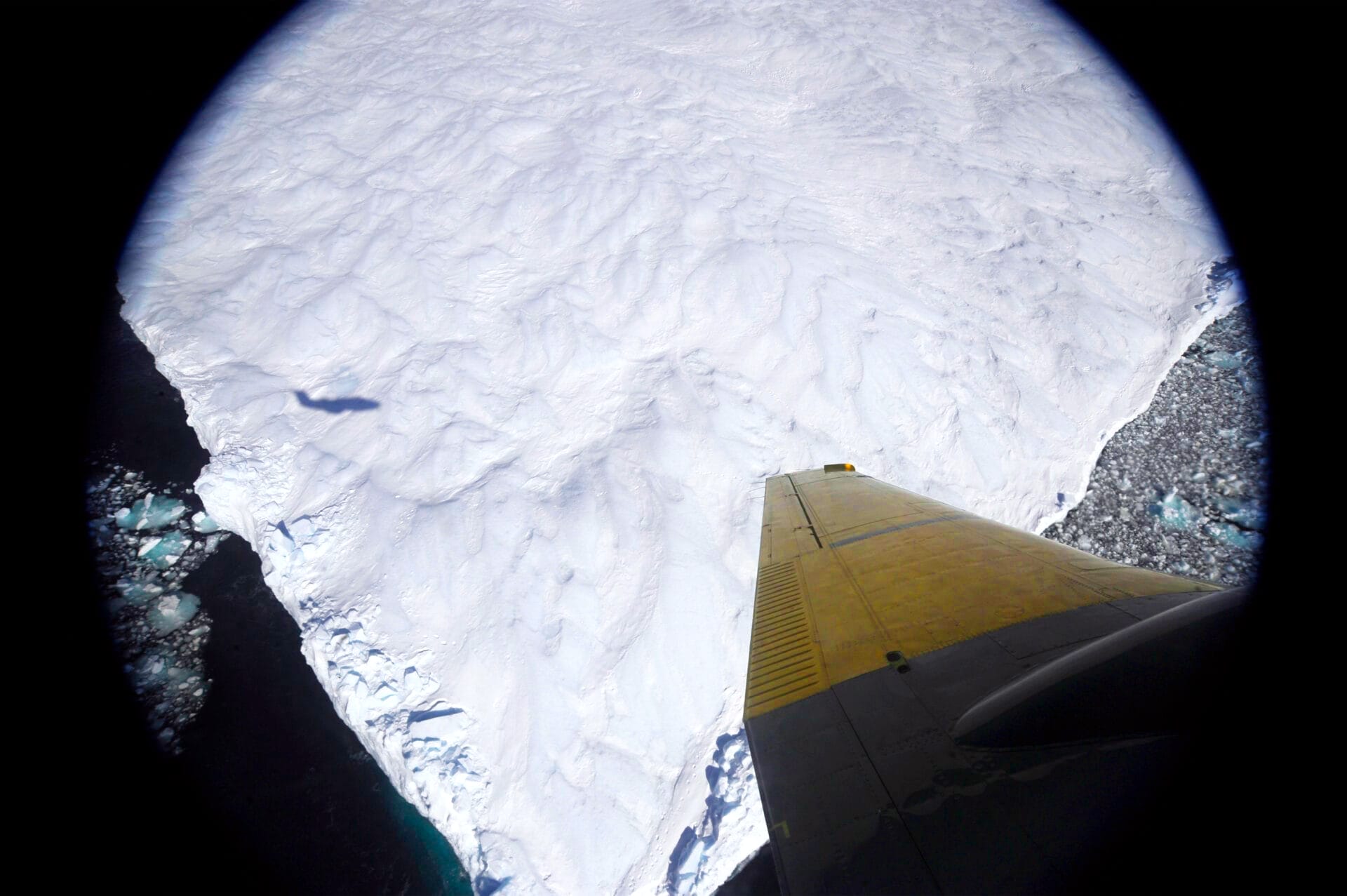 El ala amarilla de un avión se extiende sobre un vasto paisaje cubierto de nieve, contribuyendo a la vigilancia medioambiental, mientras la sombra del avión se proyecta sobre el hielo que hay debajo y el agua oscura salpicada de trozos de hielo marca el límite.