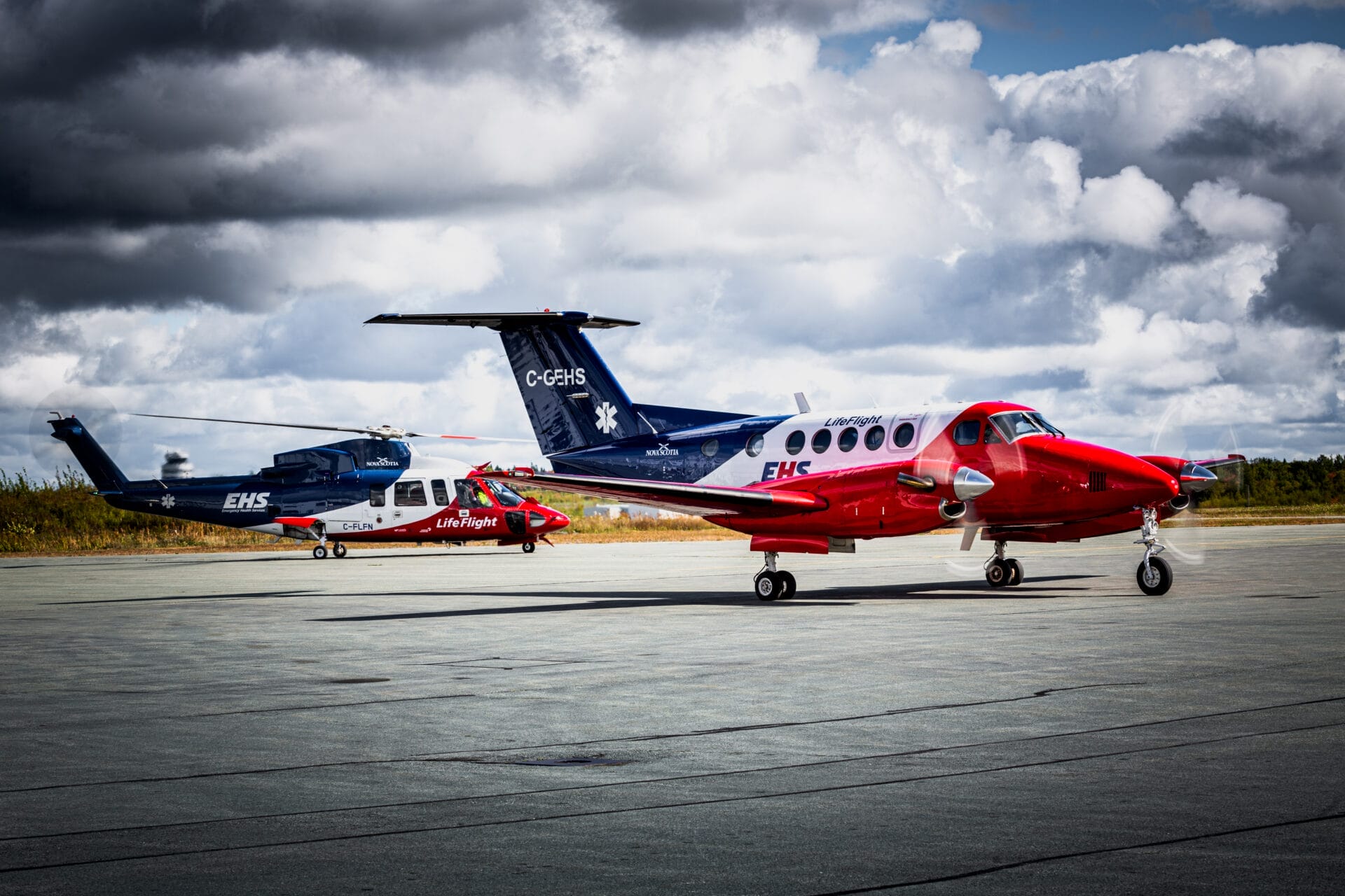 A red and white Air Ambulance plane and a blue and white medical helicopter are parked on an airport runway under a cloudy sky, with grassy fields stretching into the background.