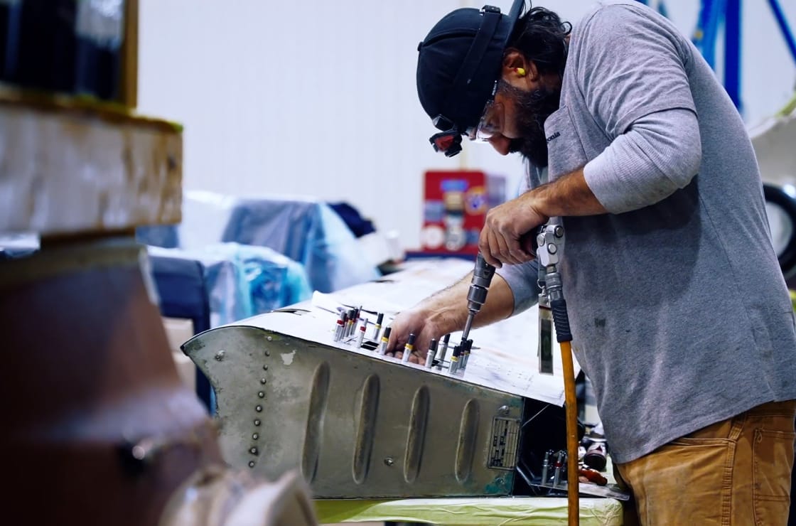 A man wearing safety goggles and a cap uses a power tool to work on a metal aircraft part in a workshop, surrounded by equipment and materials.