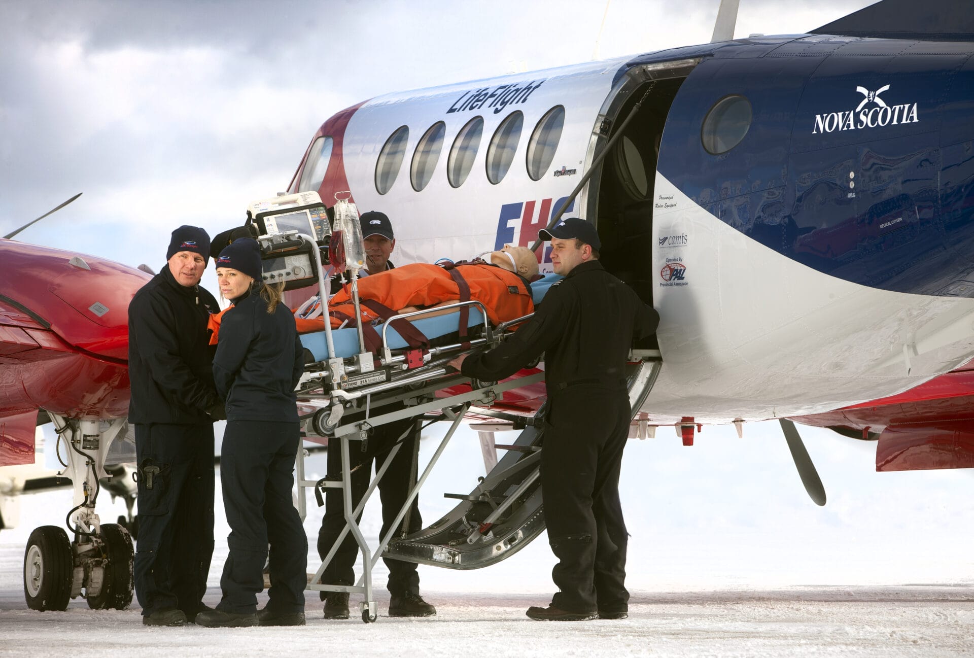 Four paramedics load a patient on a stretcher into a Nova Scotia LifeFlight Air Ambulance on a snowy surface, preparing for air transport under cloudy skies.