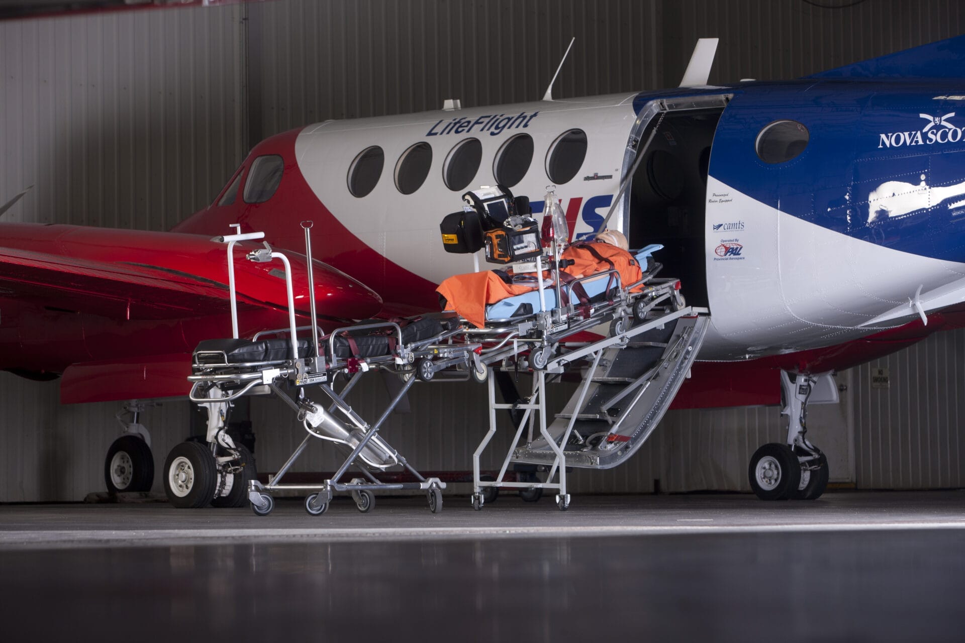 A medical stretcher is positioned on a ramp leading into an Air Ambulance LifeFlight plane parked in a hangar, ready for patient transport.