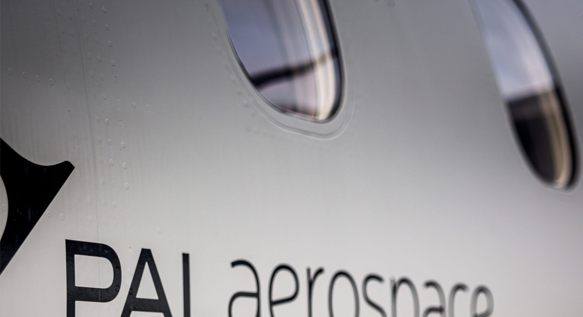 Close-up of an aircraft fuselage showing two oval windows and part of the PAL aerospace logo on the side, with light reflecting off the surface.