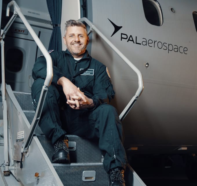 A man in a black jumpsuit sits smiling on the stairs of an aircraft with PAL Aerospace written on the side. The aircraft door is open, and the setting appears to be inside a hangar or on an airport tarmac.
