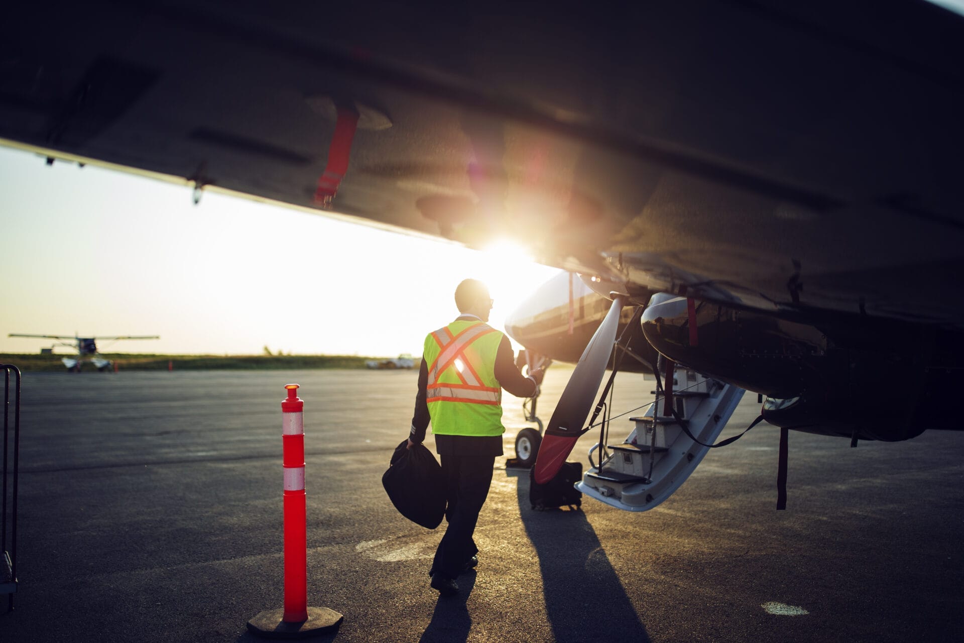 A person in a reflective vest carries a bag while walking toward the steps of an airplane at sunset, with sunlight shining through the scene on an airport tarmac.