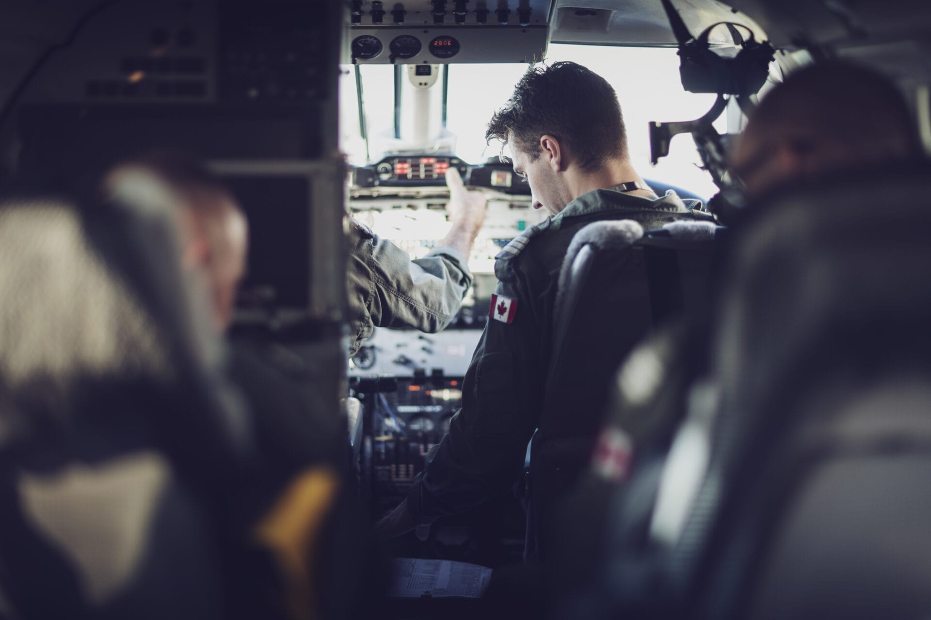 Two pilots sit in the cockpit of an aircraft, focused on the controls and instruments. The image is taken from behind, showing the pilots uniforms and equipment inside the cockpit.