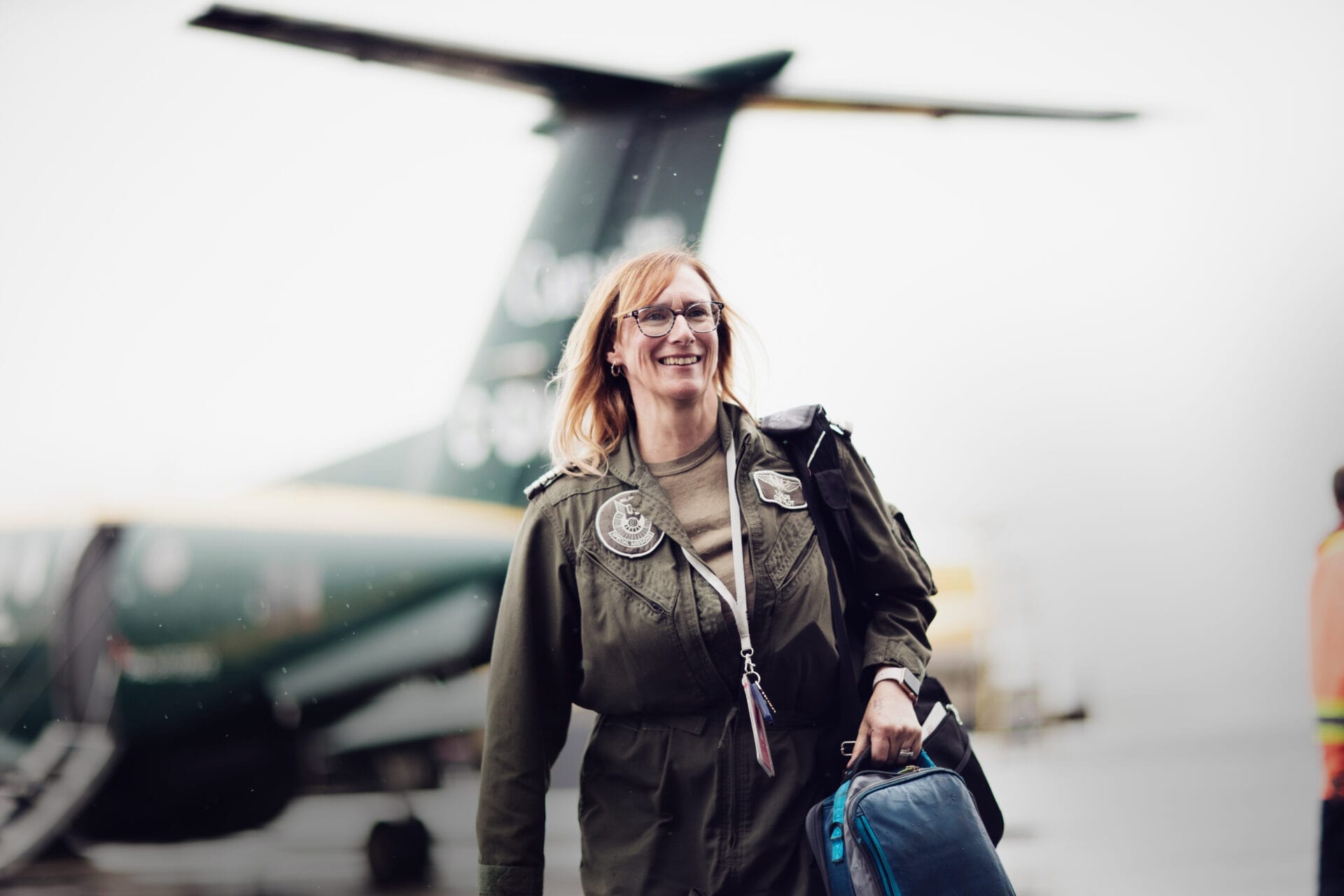 A woman in a green pilots uniform smiles while walking on an airport tarmac, carrying a blue bag over one shoulder. A small aircraft and another person in a safety vest are visible in the background.