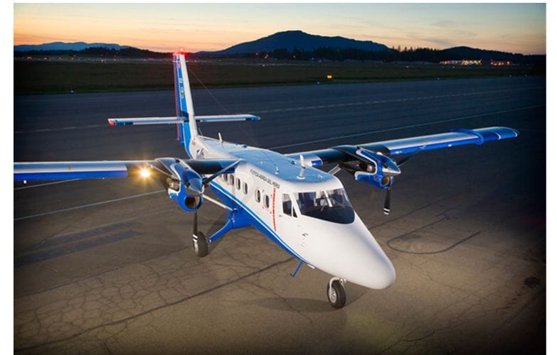 A white and blue twin-engine propeller airplane with nose-mounted equipment is parked on an airport runway at dusk, its lights glowing as mountains rise in the background.