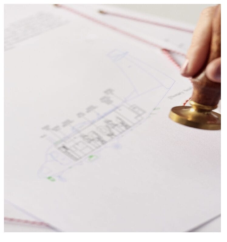 A hand presses a round brass stamp onto a sheet of paper with a technical drawing or blueprint and text, indicating document approval or certification.