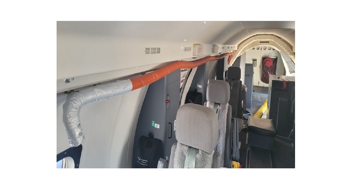 The interior of an aircraft with exposed air conditioning ductwork, gray passenger seats, and overhead compartments; an orange and silver pipe runs along the wall, and yellow tubing is visible near the rear, indicating auxiliary air conditioning features.