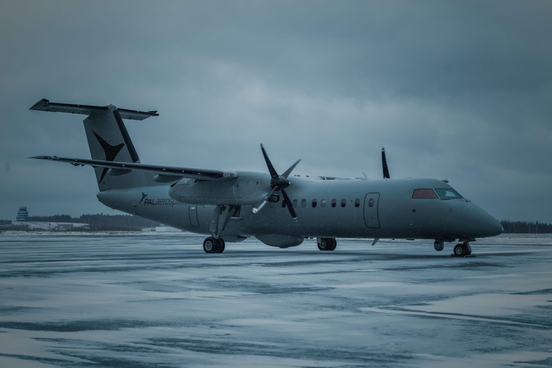 A gray twin-engine turboprop airplane, a true force multiplier, sits on a snow-covered runway under a cloudy, overcast sky, with a few distant buildings visible on the left side of the image.