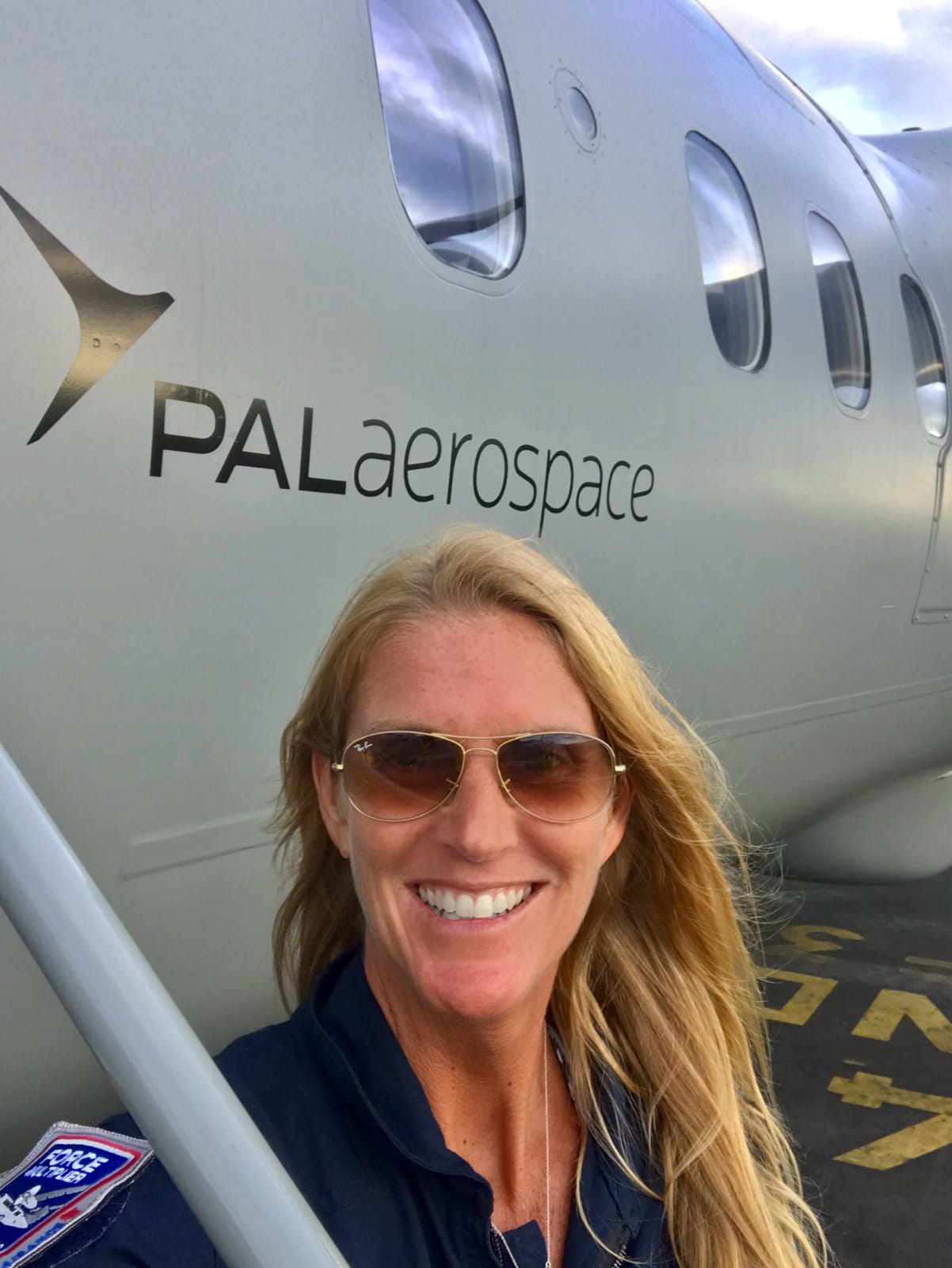 A smiling woman with long blonde hair and sunglasses takes a selfie in front of a grey PAL Aerospace aircraft. Wearing a dark aviation uniform, she stands on an airfield—ready to be a true force multiplier in aviation.