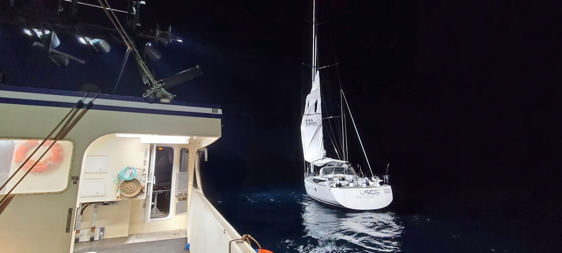 A sailboat with white sails, illuminated on dark water at night, floats near the well-lit deck of a larger vessel—casting an air of yacht mystery over the pitch-black sea and sky.