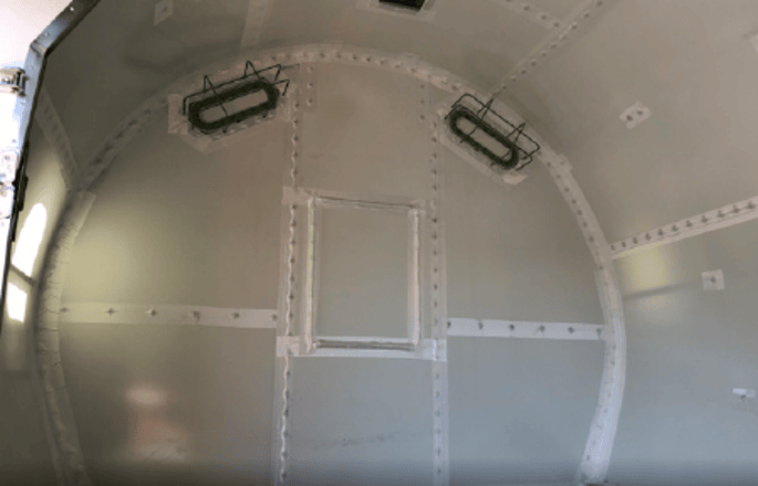The interior of a metal, cylindrical structure features curved walls with visible bolts, two rectangular lights on the ceiling, and a square panel in the center of the back wall—showcasing subtle interior modifications.