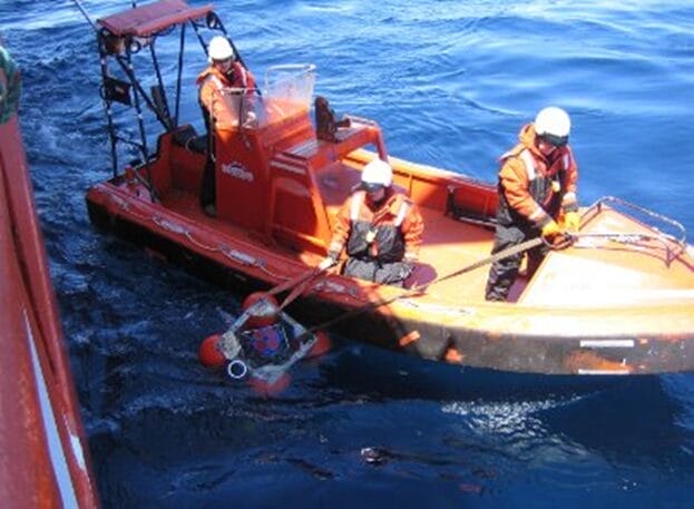 Tres personas vestidas con ropa de seguridad naranja y cascos se encuentran en una lancha de rescate naranja, utilizando una pértiga para sacar a una persona del agua y ponerla a salvo cerca de una embarcación más grande, lo que forma parte de las soluciones de predicción esenciales para la seguridad marítima en medio de las olas azules.