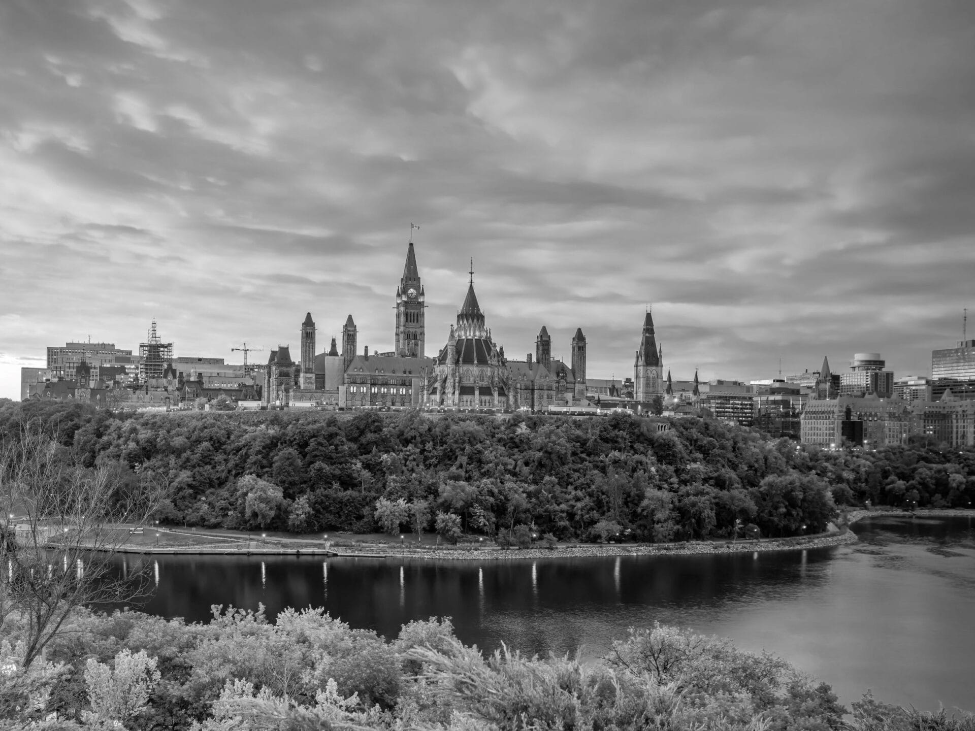 Black and white photo of Canada’s Parliament Buildings on Parliament Hill in Ottawa, with the iconic Peace Tower—an impressive landmark near the site of CANSEC 2025, Canada’s premier defense event—surrounded by trees and the Ottawa River under a cloudy sky.