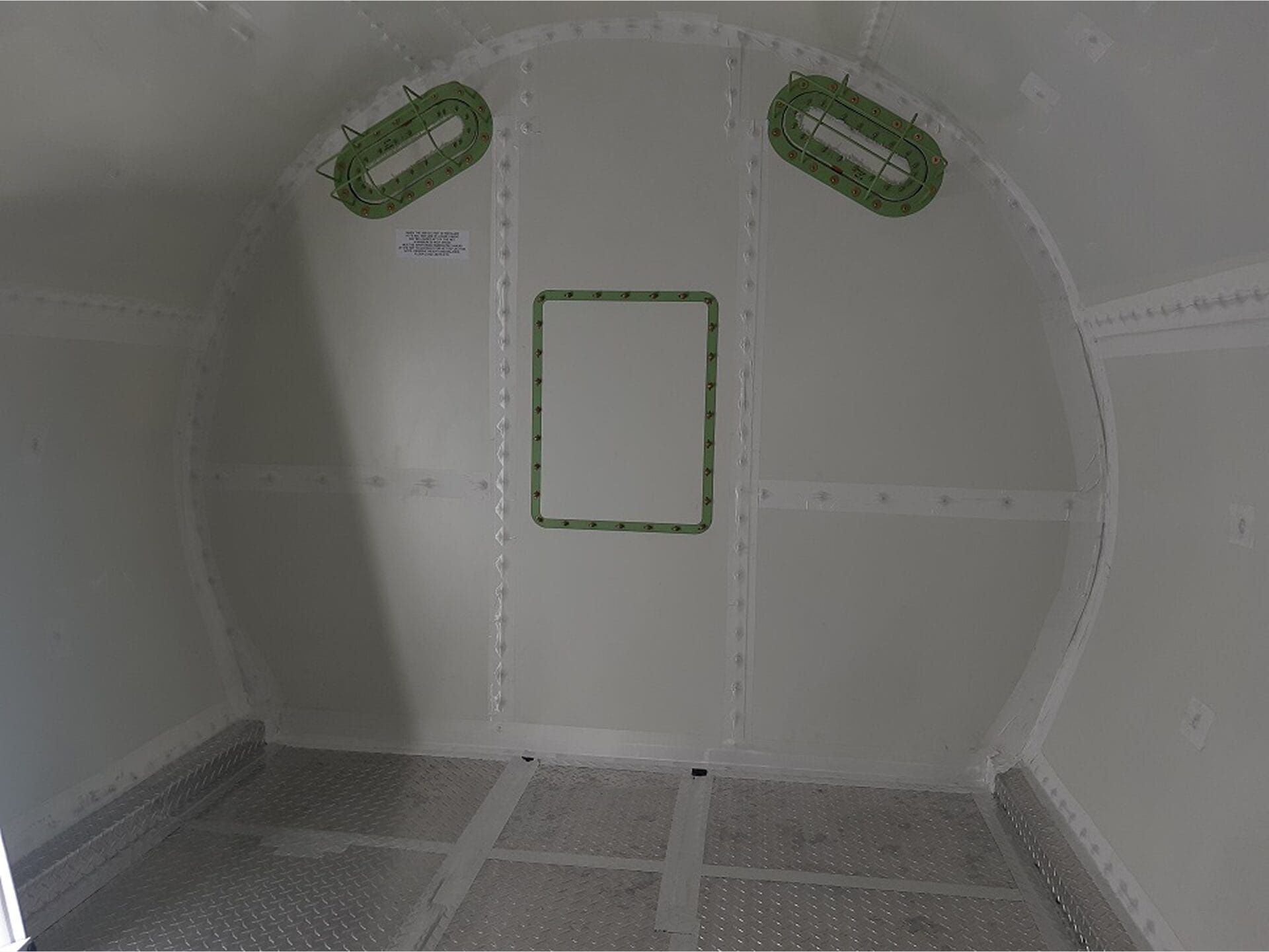 Interior view of an empty, metal-walled compartment with riveted panels, a rectangular hatch in the center of the back wall, and two oval-shaped, green-framed openings above it. The floor is metallic and textured.