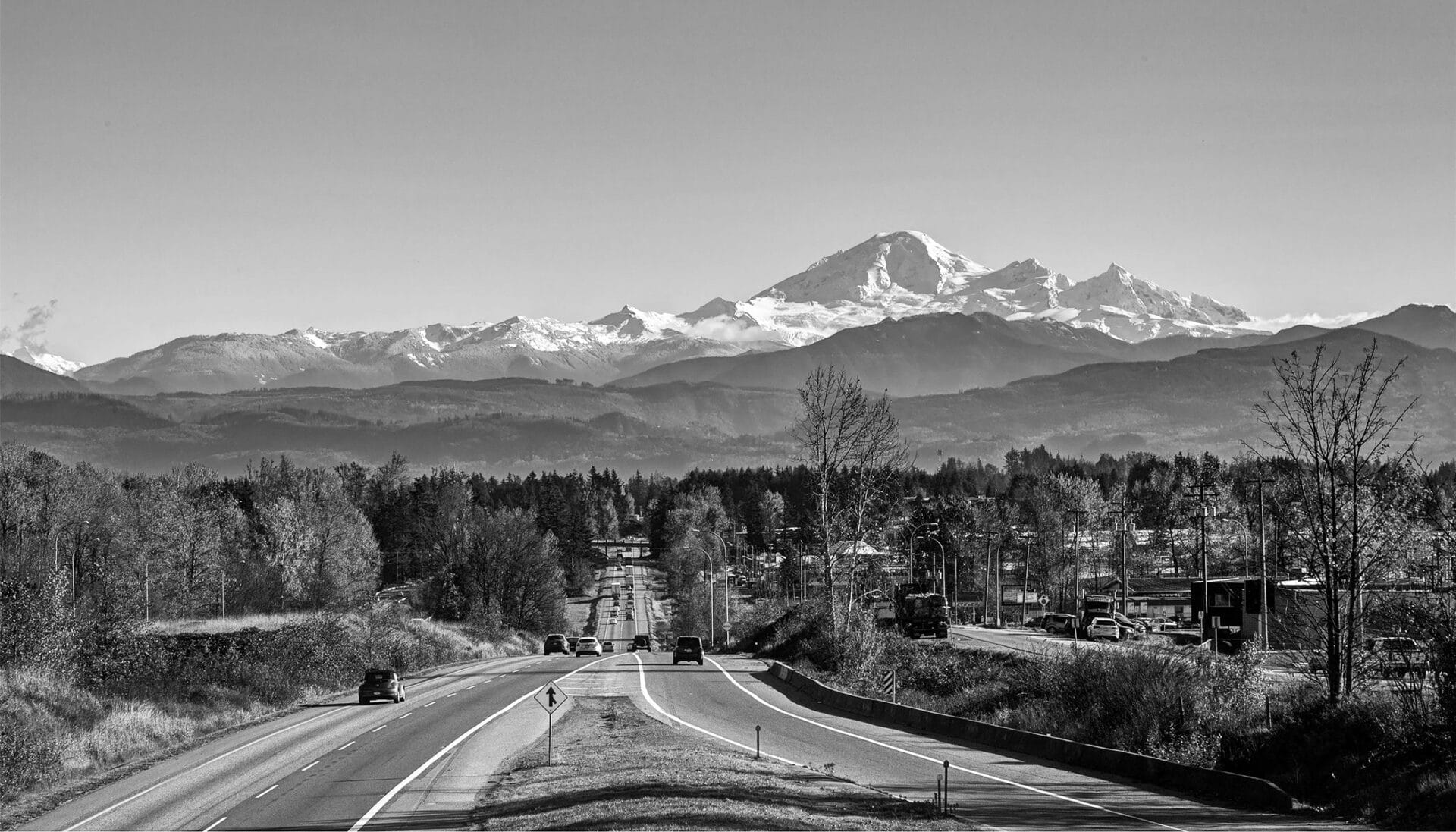 A scenic road leads toward snow-capped mountains under a clear sky, with cars driving and trees lining both sides. Captured in black and white, the landscape's contrast and detail evoke the precision of aerospace engineering.