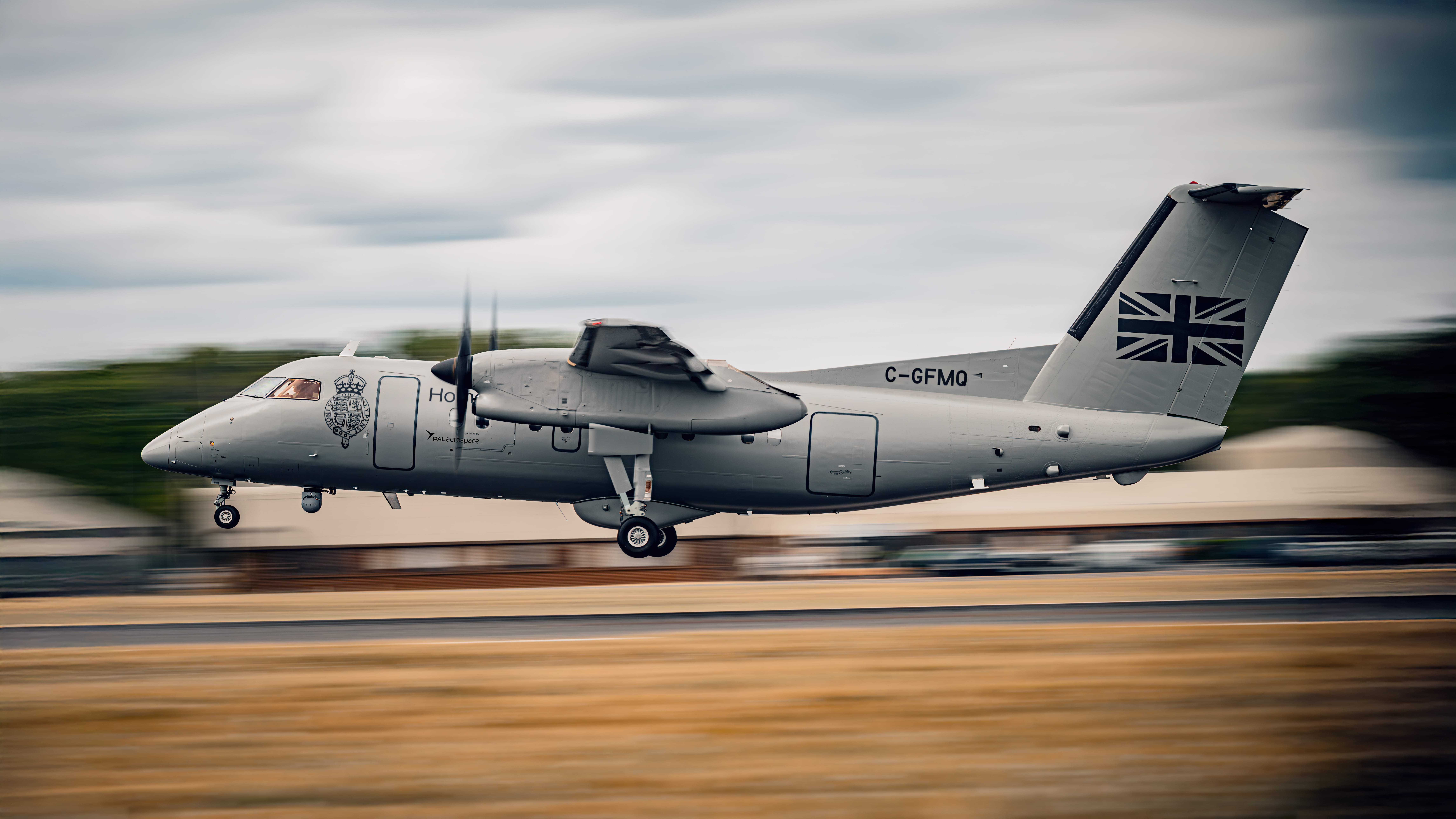 A grey military aircraft with a Union Jack on the tail and UK Home Office registration C-GFMQ takes off from a runway. The blurred background, with buildings and trees, emphasizes its swift motion.