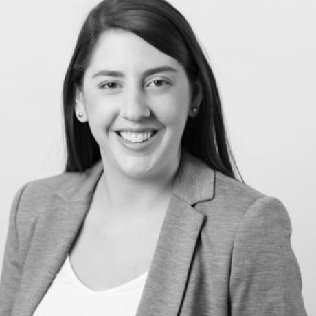 Portrait en noir et blanc d'une femme aux longs cheveux bruns, vêtue d'un blazer clair sur un haut blanc, souriant à l'appareil photo devant un fond uni — idéal pour votre prochaine discussion informelle ou votre prochaine réunion de planification.