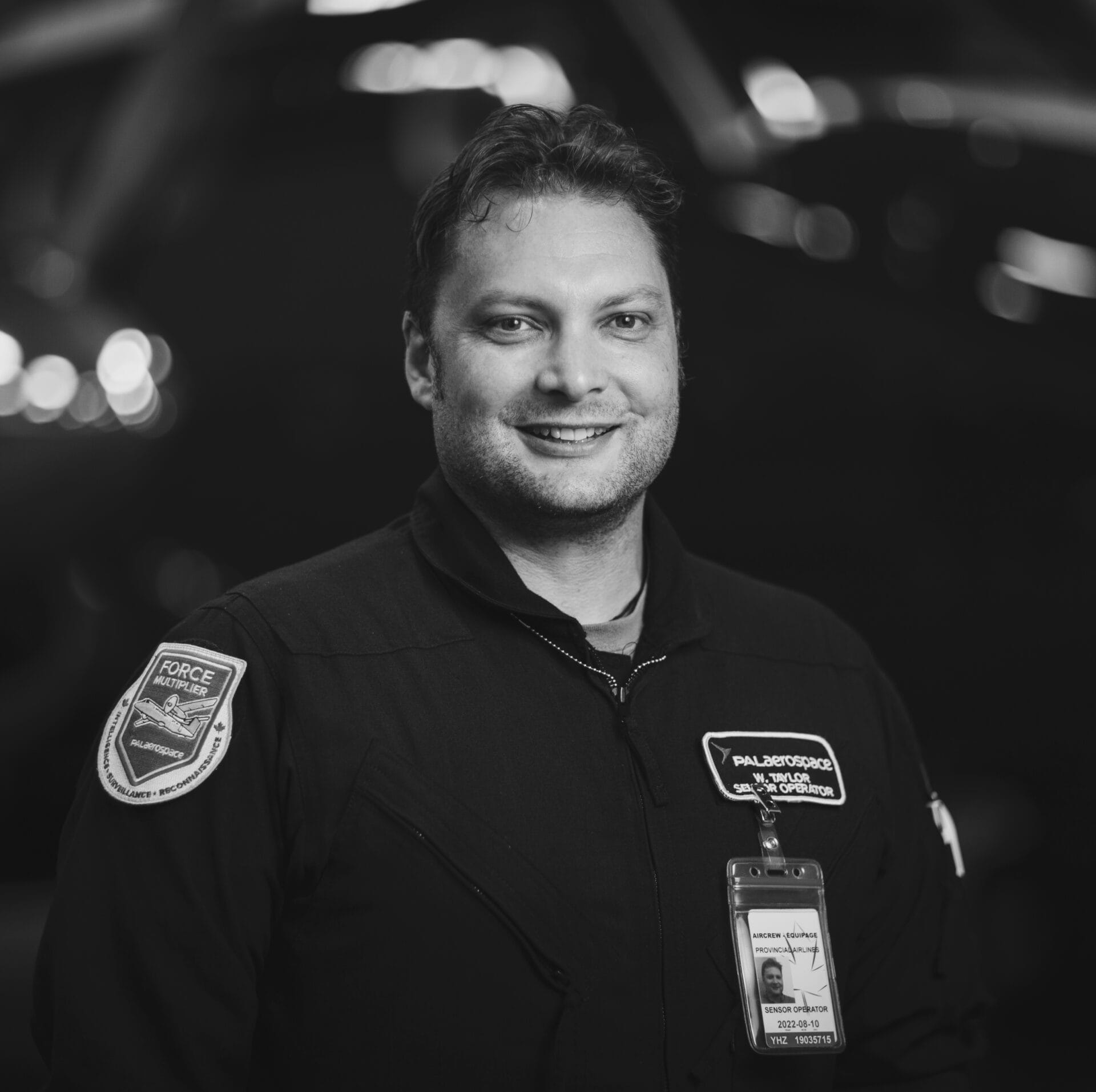 Un homme vêtu d'un uniforme sombre, arborant un écusson « PALOS VERDES-PSD CHOPPER » et un badge d'identification, sourit à la caméra, debout devant un hélicoptère flou. Cette photo en noir et blanc rend bien l'esprit de la préparation d'une mission.