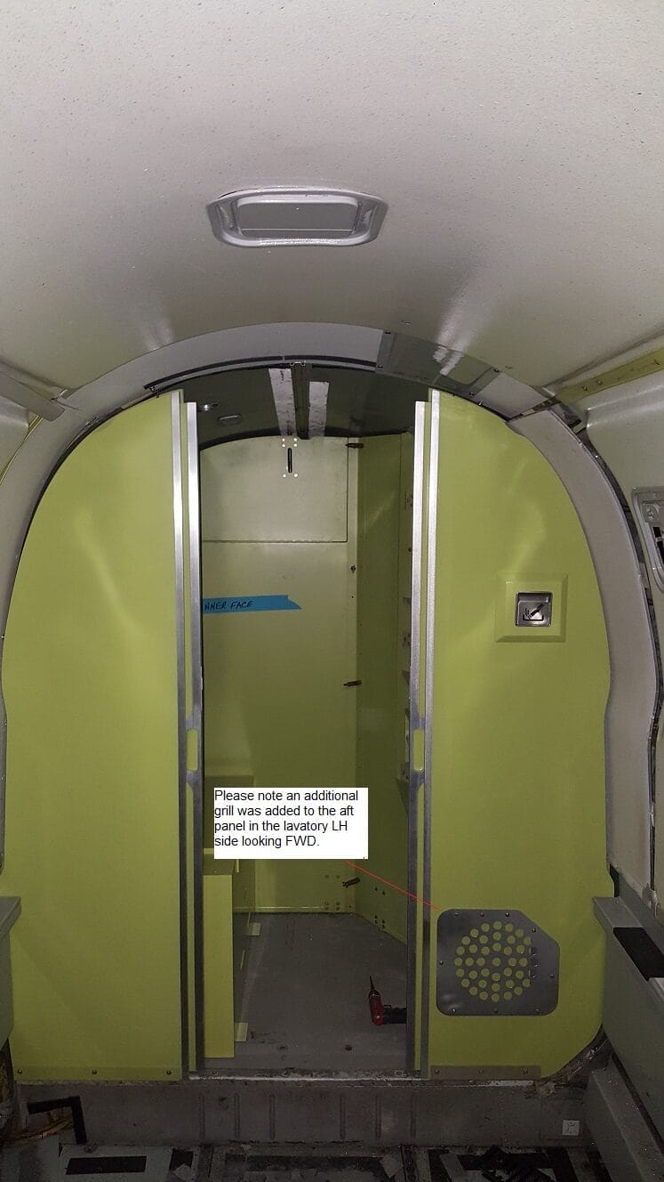 Interior of an aircraft with greenish panels, a partially open door, exposed floor, and a sign on the door reading, “Please note an additional panel in the lavatory LH side looking FWD,” likely related to lavatory installation or bathroom plumbing.