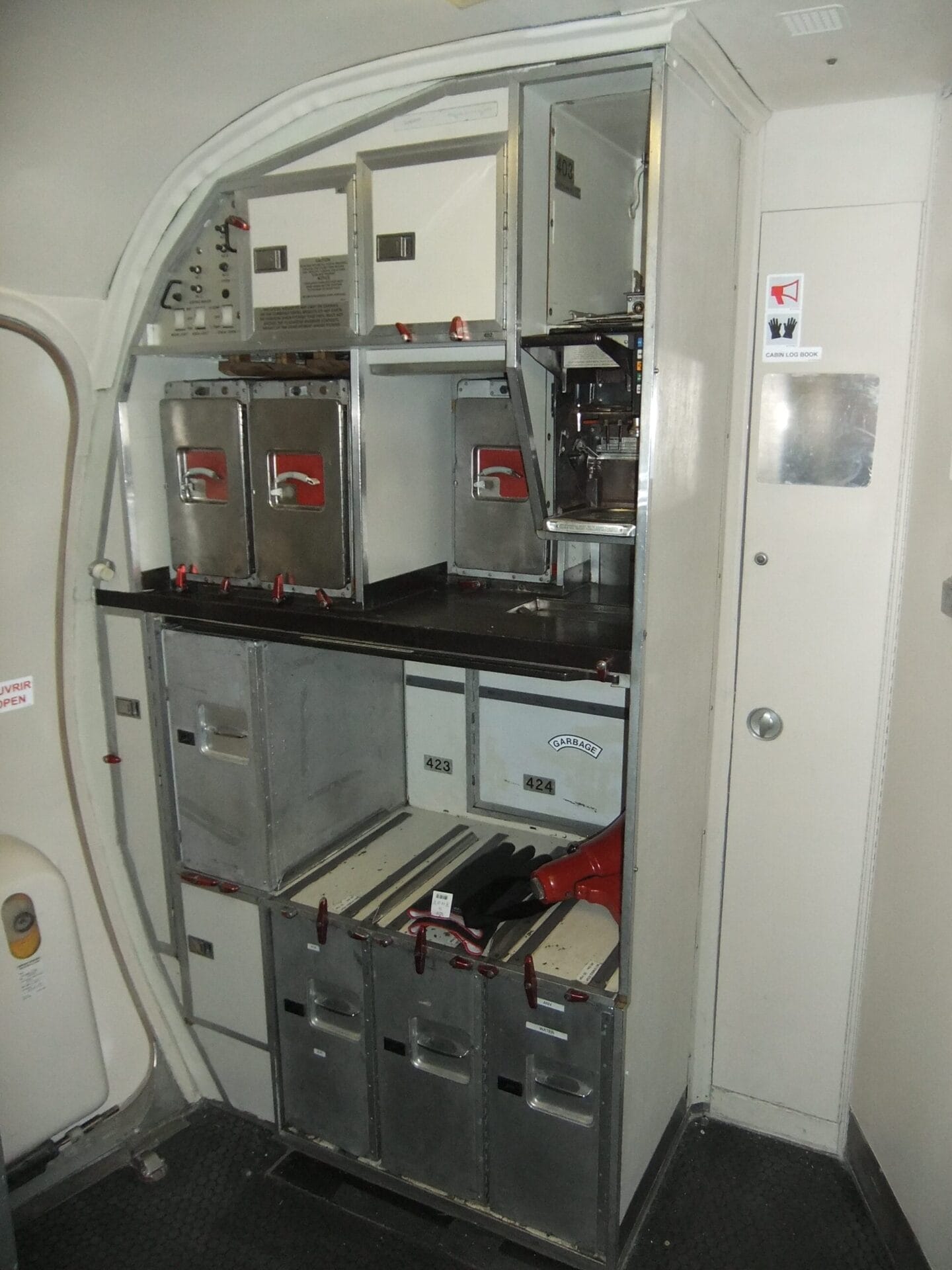 Airplane galley with metal storage compartments, ovens, and drawers for food and drink service, located near an aircraft door. This multiple galley installation features metallic surfaces with red latches and various labels.