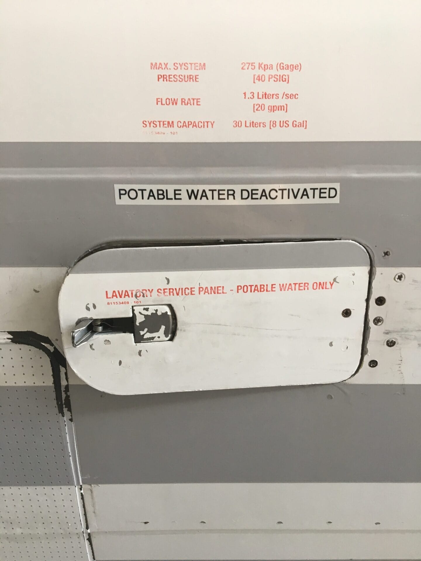Close-up of an aircraft lavatory service panel with labels stating POTABLE WATER DEACTIVATED and specifying potable water system pressure, flow rate, and capacity details above the panel.