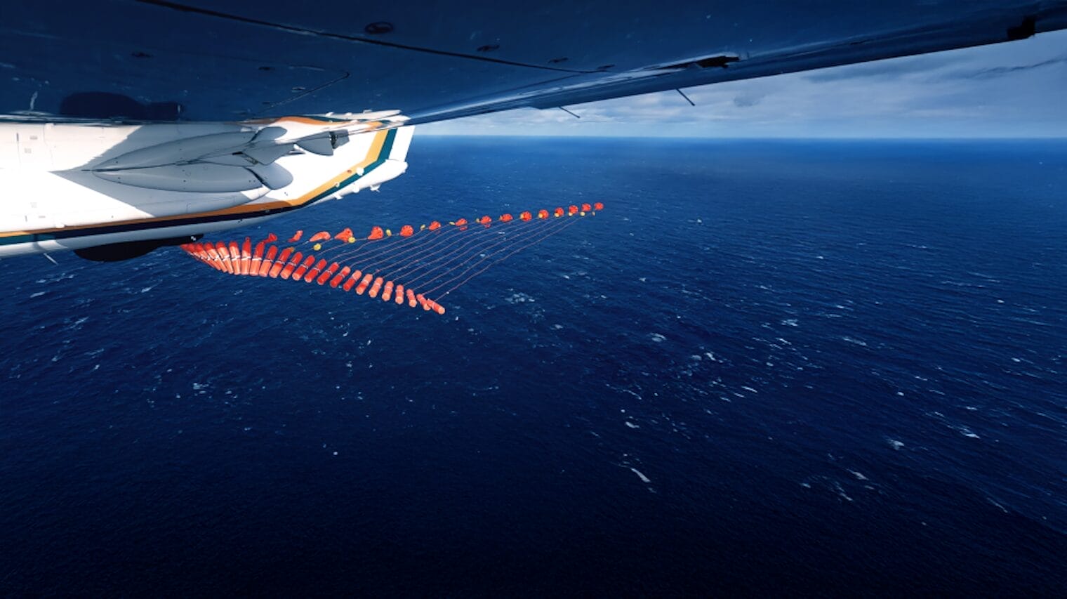 A view from under an airplane wing shows a sequence of red objects, likely engineering devices to save lives, being deployed over the open ocean from the sky beneath a cloudy expanse.
