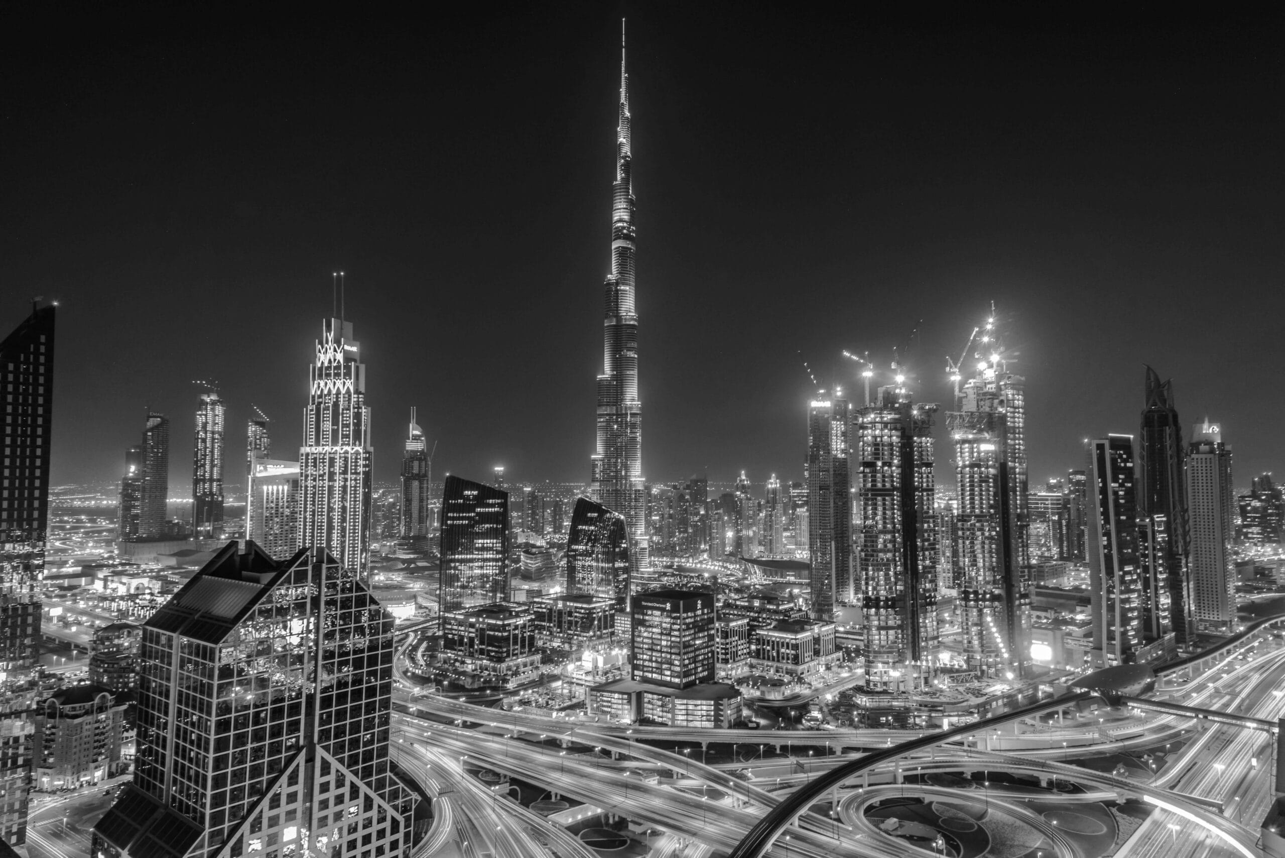 A black and white nighttime cityscape of Dubai features the illuminated Burj Khalifa towering above other skyscrapers, with busy roads weaving through the vibrant city, capturing the dynamic spirit ahead of the Dubai Airshow 2025.