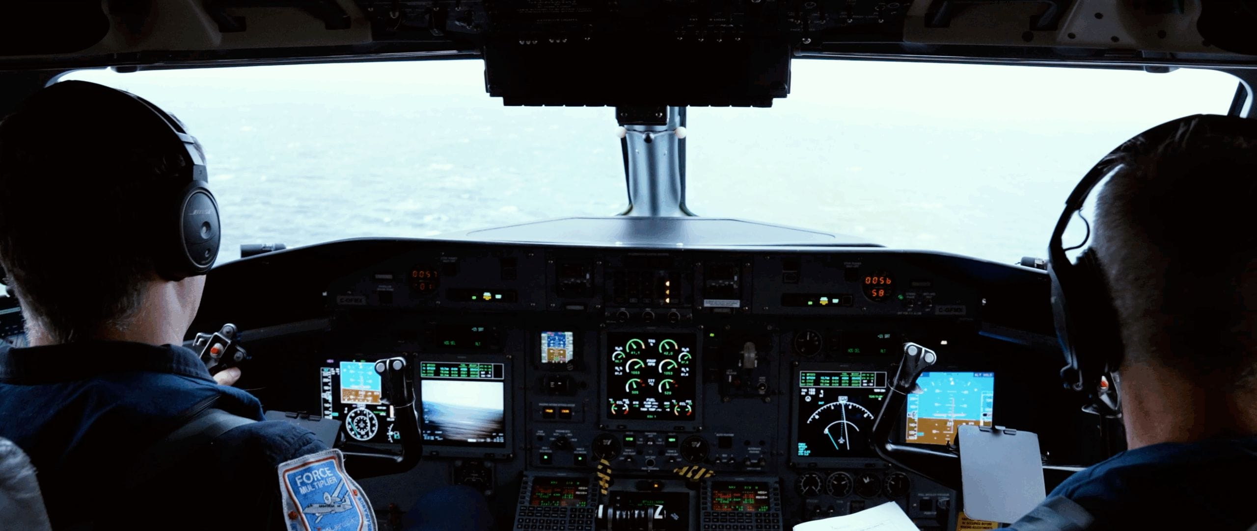 Two airline pilots sit in the cockpit of an airplane, facing a control panel filled with illuminated instruments and screens, giving you an exclusive look as they navigate through a cloudy sky.