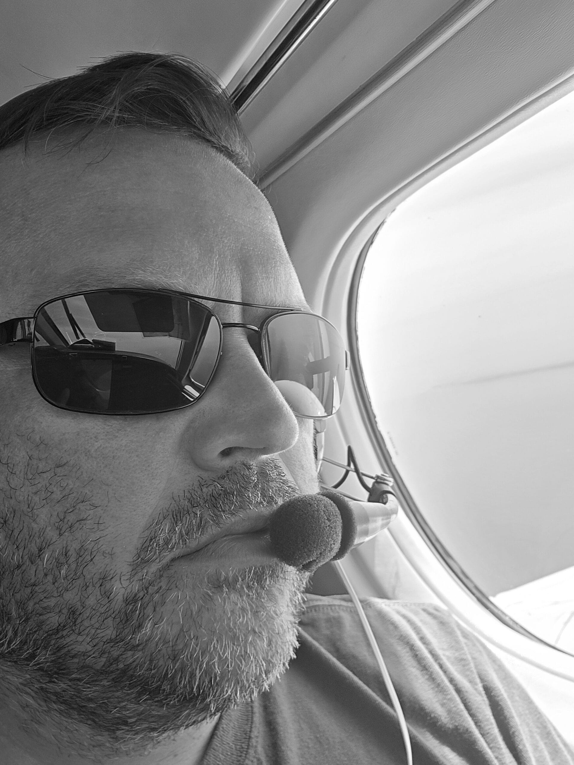 Un hombre con gafas de sol y un micrófono de diadema está sentado junto a la ventanilla de un avión, mirando hacia fuera. Esta fotografía en blanco y negro ofrece una visión exclusiva del mundo de los pilotos de línea aérea en misiones especiales.
