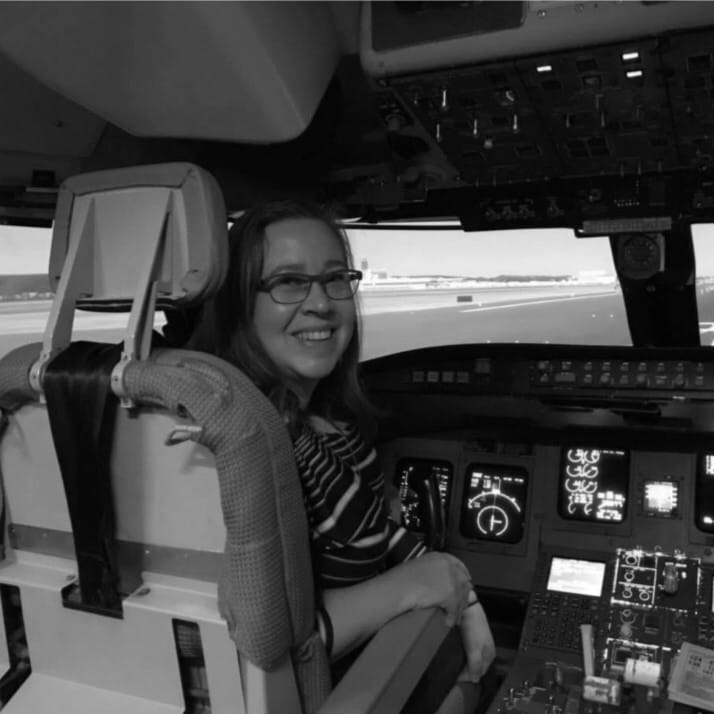 Una mujer con gafas y una camiseta a rayas está sentada en la cabina de un avión, sonriendo a la cámara. Los mandos de vuelo y los paneles de instrumentos la rodean, lo que refleja el papel fundamental que desempeñan los pilotos en las aerolíneas cada día.