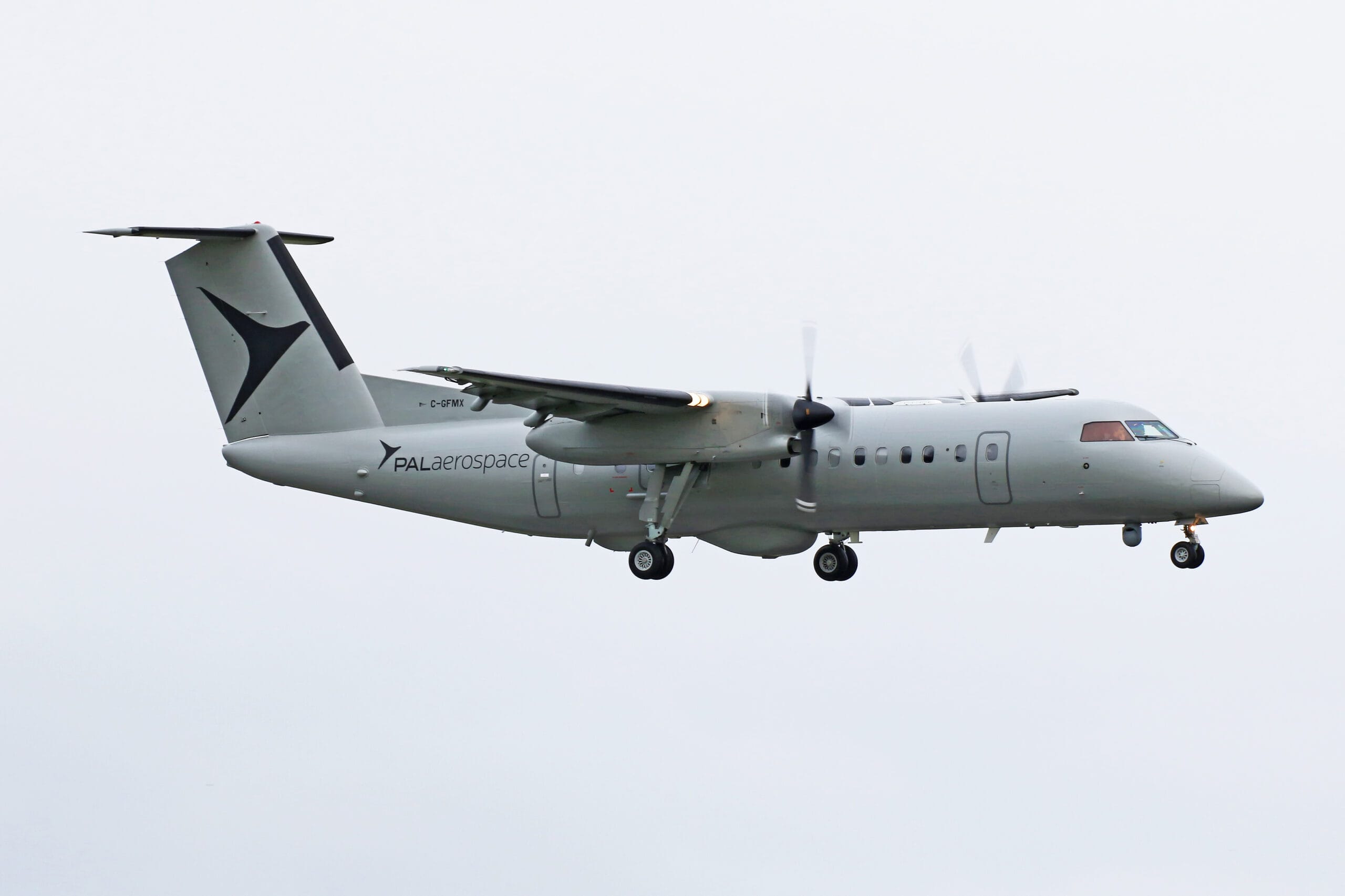 A light gray De Havilland PAL Aerospace propeller airplane is flying against an overcast sky, with its landing gear extended and propellers spinning, resembling a broadcast of aviation excellence in motion.